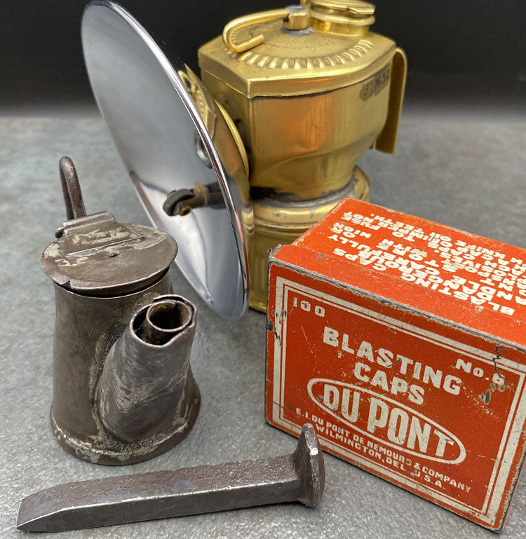 Collage of mining tools including box of blasting caps, a metal spike, a brass headlamp and a metal pot.