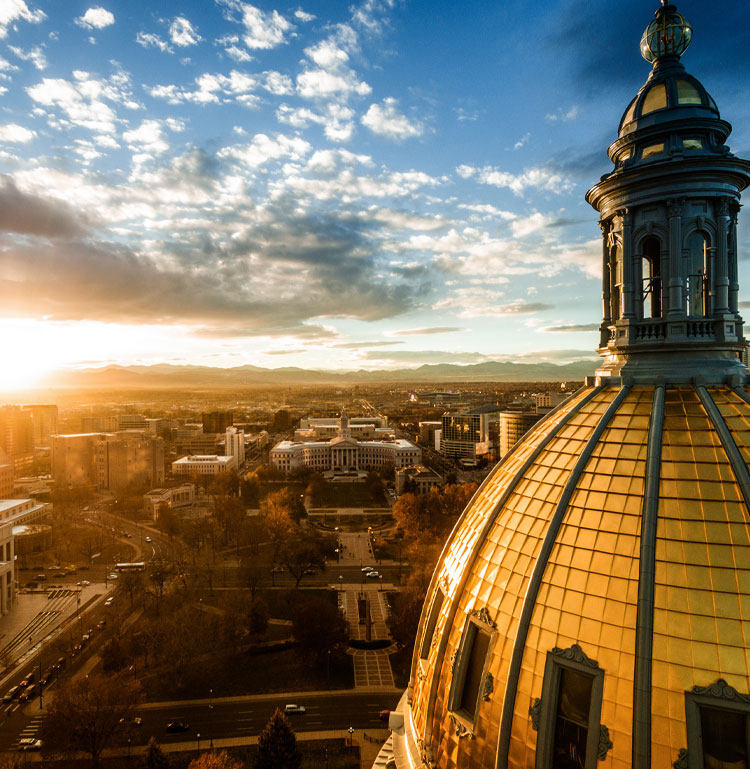 Drone shot of Capitol dome in Denver, looking west. In distance the city and county building, mountains and sunset.