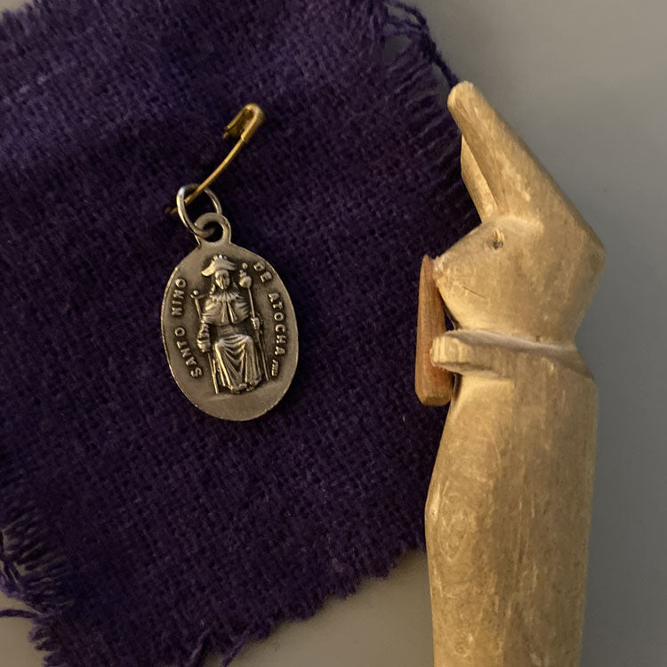 Small medal with embossed image of Santo Nino De Atocha pinner to burlap and wooden carven rabbit holding a carrot.