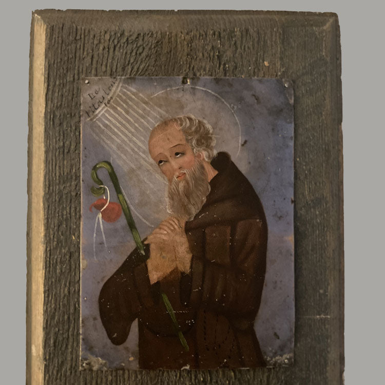 retablo, with image of man praying while light shines down on him. He is holding a staff with a small bag on the end. the image is mounted on a rough wooden plaque.