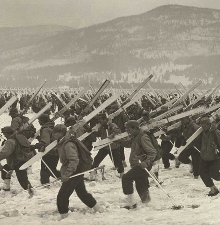 Black and white photograph of a group of soldiers trudge through snow while carrying skis.