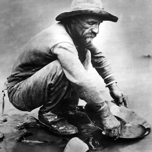 Miner wearing large brimmed hat squatted by stream panning for gold. 