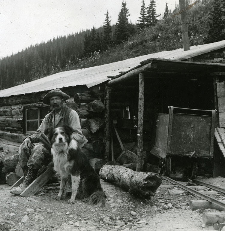 Photo of miner and dog sitting out front of log mine entrance. An ore cart is nearby.